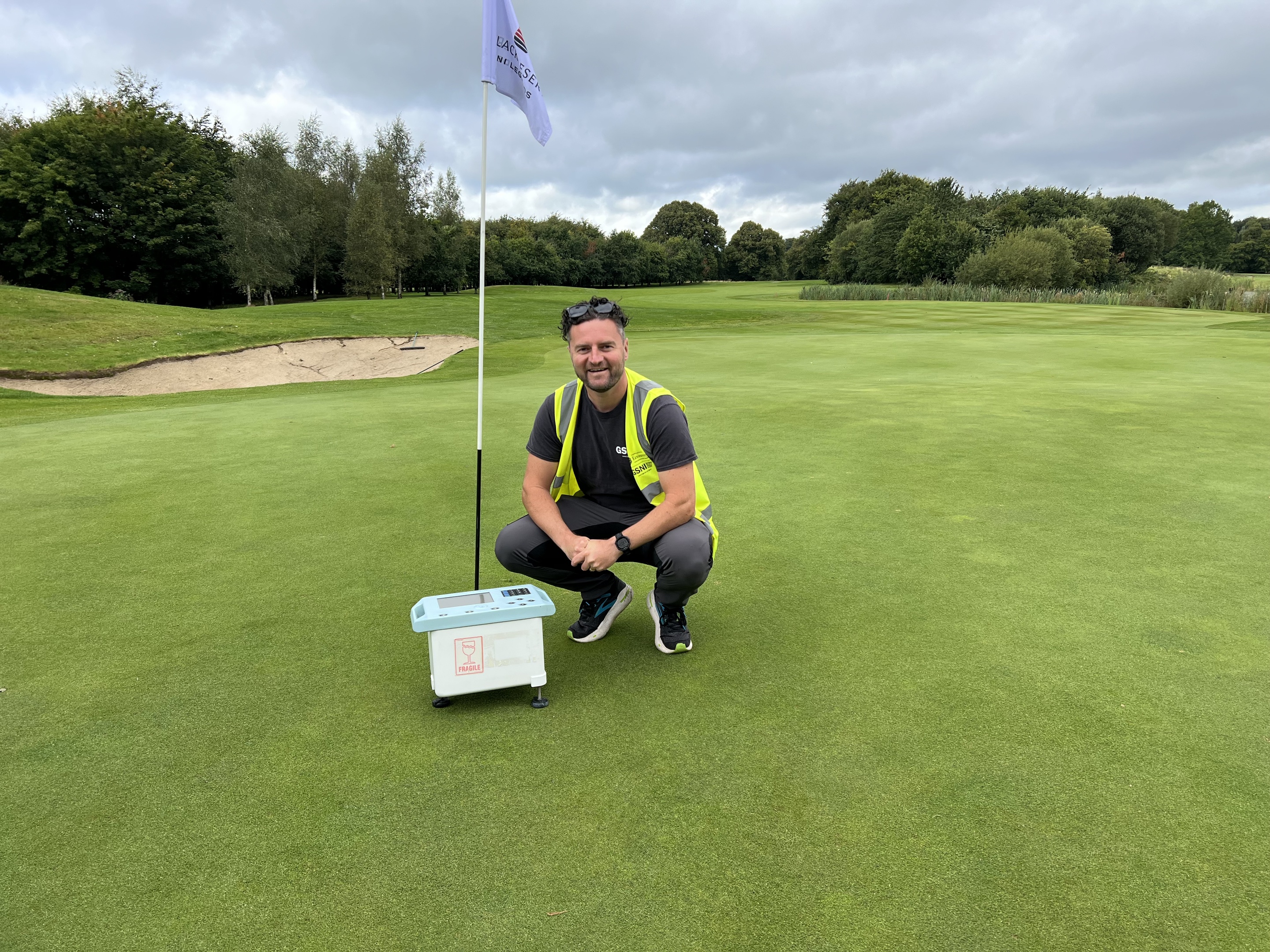 A male researcher crouches alongside a gravity survey instrument positioned on a putting green, with a fairway and sandpit beyond