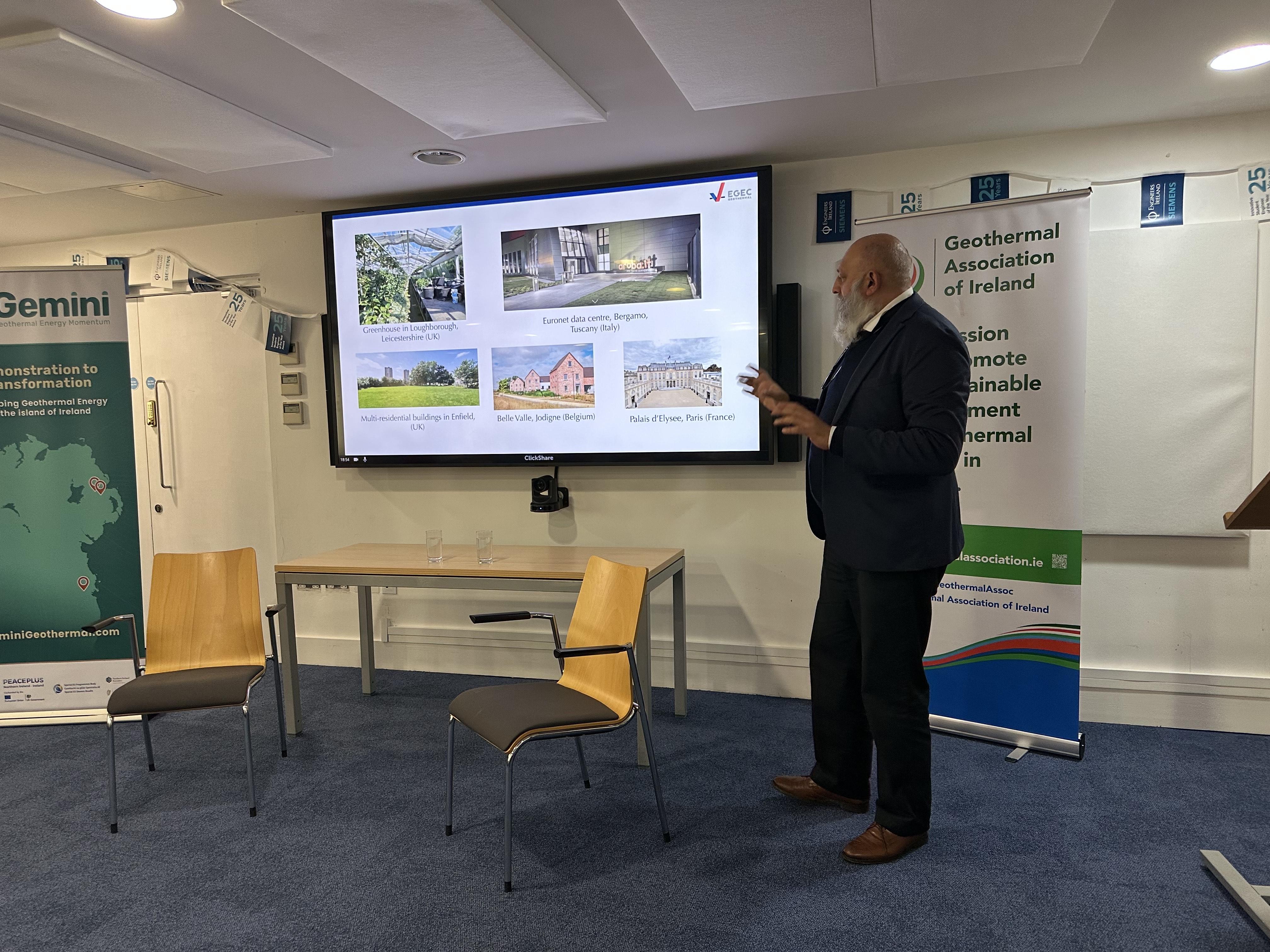 Sanjeev Kumar is talking animatedly in front of a large screen featuring buildings in Europe: residential buildings in Enfield, UK; Belle Valle, Jodigne (Belgium); Palais d'Elysee, Paris (France); Euronet data centre, Bergamo (Italy).