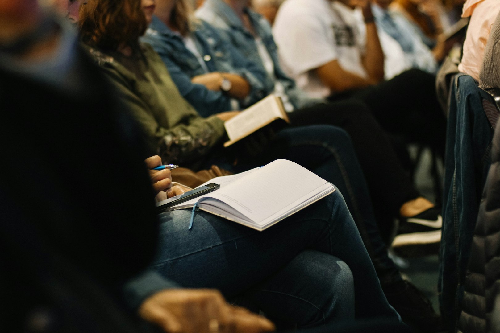 A row of seated people with focus upon the pads and pens on their laps, for taking notes